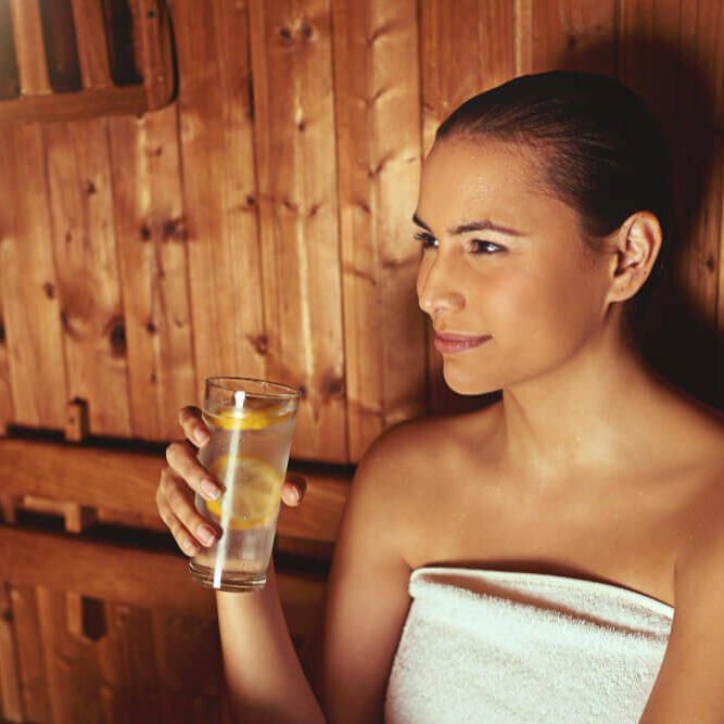 Femme dans un sauna et verre d'eau dans un spa pour réfléchir à un traitement de sudation ou de détoxification pendant ses vacances dans un complexe hôtelier de luxe, personne heureuse et détendue pendant ses vacances dans un lodge, dans un hammam pour soulager le stress cutané ou réfléchir.